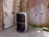 A metal barrel converted into a shelf unit, placed in an outdoor area with a brick wall and a weathered door. Anteprima dell’immagine prodotto