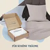 A cardboard box with bedding inside is placed on a bed with pillows and sheets. Produktbild-Vorschau