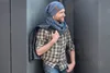 A man leaning against a dark wall, wearing a blue knit hat and scarf, and a plaid shirt. Produktbild-Vorschau