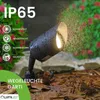 A black outdoor spotlight is shown in a garden setting, with water droplets on it, suggesting it is waterproof. The background includes plants and mulch. Náhľad obrázku produktu