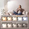 A woman sits on a couch holding a mug, with a floor lamp in the foreground. The image displays different color temperatures and dimming levels of the lamp. Produktbild-Vorschau