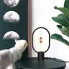 A modern magnetic balance lamp is placed on a dark table next to a framed artwork of moon phases and a fiddle-leaf fig plant. A white cat is looking at the artwork. Produktbild-Vorschau
