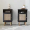Two black nightstands with rattan fronts and wooden legs are displayed against a white wall. Anteprima dell’immagine prodotto