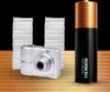 A digital camera and a large Duracell battery stand next to stacks of paper on a wooden surface, with a black background. Náhled obrázku produktu