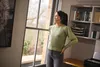 A woman wearing a headset stands by a window, looking out. She is in a home office setting with shelves and a printer visible. Produktbild-Vorschau