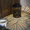 A black metal lantern with a leaf pattern is placed on a stone surface next to a cup of coffee. The lantern casts intricate leaf-shaped shadows on the surface and the wooden wall behind it. Produktbild-Vorschau