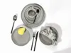 A collection of grey ceramic plates, bowls, and a cup, with a spoon, fork, knife, and a pear. Produktbild-Vorschau