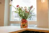 A colorful vase filled with flowers sits on a countertop in front of a window with a city view. Produktbild-Vorschau