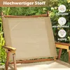 A close-up of a beige camping chair with wooden armrests, set outdoors with green foliage and yellow flowers in the background. Text overlays highlight product features. Produktbild-Vorschau