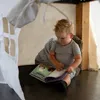 A young child is sitting under a table, reading a book inside a tent. Produktbild-Vorschau