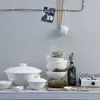 A collection of white ceramic tableware, including bowls and a large lidded serving dish, arranged on a shelf next to a small portable stove and a bottle of soy sauce. A string of twine hangs from a rod above, with a photograph and chopsticks. Produktbild-Vorschau