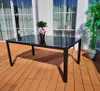 A black glass-topped table with a metal frame sits on a wooden deck, with a potted plant to the right and windows in the background. Produktbild-Vorschau
