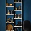 A white shelving unit filled with various kitchen storage items against a dark blue wall. Produktbild-Vorschau