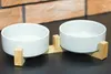A white ceramic double pet bowl set on a wooden stand, placed on a dark surface with a brick wall in the background. Produktbild-Vorschau