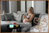 A pregnant woman is sitting on a couch, looking at her phone. In front of her is a small table with a diffuser, fruit, and a glass of juice. Produktbild-Vorschau