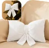 A white bow-shaped pillow is placed on a beige sofa. A small inset image shows a similar bow on a Christmas tree. Produktbild-Vorschau