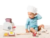 A baby wearing a chef's hat sits on a rug next to a toy toaster and breakfast accessories. Anteprima dell’immagine prodotto