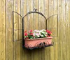 A black metal window-style flower holder with pink and red flowers is mounted on a weathered wooden fence. Produktbild-Vorschau