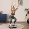 A woman is performing a squat exercise on a vibration plate in a living room setting. Náhled obrázku produktu