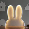 A bunny-shaped night light with a white outline and warm yellow light illuminating the interior, placed on a wooden surface against a gray wall with other wooden cutouts of a house, a car, and a hedgehog. Produktbild-Vorschau