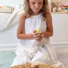 A young girl in a white dress is sitting on the floor, holding a small colorful toy in her hands. There are some wooden discs stacked on a woven mat in front of her. Anteprima dell’immagine prodotto