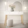 A white vanity table with wooden legs, a vase of flowers, and makeup on top, in a well-lit room with decorative wall molding and a floor lamp. Produktbild-Vorschau