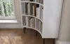 A white, curved bookshelf filled with books, standing on black legs next to a window. Produktbild-Vorschau