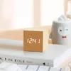 A wooden cube alarm clock with a digital display sits on a stack of books next to a ceramic penguin mug. Produktbild-Vorschau