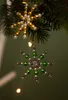 Two beaded snowflake ornaments hanging from a pine branch, with blurred lights in the background. Produktbild-Vorschau