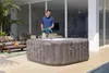 A man is filling an inflatable hot tub with water from a hose in an outdoor patio area. Produktbild-Vorschau