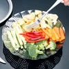 A stainless steel steamer basket filled with various cut vegetables, including potatoes, carrots, broccoli, cucumber, and red bell pepper, with a fork in the basket. Produktbild-Vorschau