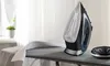 A cordless steam iron is placed on an ironing board next to a white shirt. A plant is visible in the background. Produktbild-Vorschau