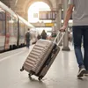 A person is pulling a champagne-colored suitcase on wheels at a train station. Náhľad obrázku produktu