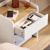 A close-up view of a white bedside table with a drawer partially open, revealing a calculator and other items. A book and a wireless charger are on the top surface. Produktbild-Vorschau