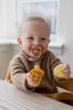 A baby sitting in a high chair, holding two pacifier toys. Produktbild-Vorschau