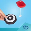A hand holding a black and white target-shaped toy, with a red plush toy hanging from a string in the background. Produktbild-Vorschau