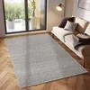 A light gray rug is placed on a wooden floor in front of a large window and a sofa.