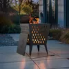 A black metal fire pit with a patterned design is lit with flames, placed on an outdoor patio with plants in the background. Náhled obrázku produktu