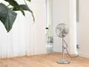 A silver stand fan is placed on a wooden floor in a room with white walls and sheer white curtains. Produktbild-Vorschau