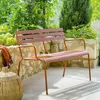 A metal garden bench with amber-colored slats and frame, placed on a patio with a stone wall and lush greenery in the background. Produktbild-Vorschau