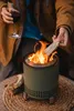 A person is holding a glass of red wine and adding a piece of wood to a small, green fire pit. Náhľad obrázku produktu