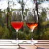 Two glasses of rosé wine are placed on a wooden table outdoors, with a blurred background of trees and foliage. Produktbild-Vorschau