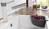 A white kitchen sink with a modern faucet and a gray colander filled with red grapes resting on the edge. Produktbild-Vorschau
