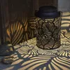 A black metal lantern with a leaf pattern is placed on a surface, casting intricate leaf-shaped shadows on the surface and a wooden wall. Produktbild-Vorschau