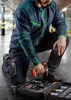 A man is kneeling in a parking garage, holding a cordless drill and working on a tool case. Produktbild-Vorschau