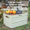 A cream-colored foldable camping box is shown outdoors on grass, with fruit and a speaker on top, suggesting its use as a table. Produktbild-Vorschau