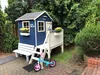 A blue wooden playhouse for children is set up in a garden, with a small bicycle in front of it. Produktbild-Vorschau