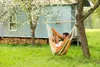 A couple relaxing in a colorful hammock hanging between two trees in a garden, with a light blue wooden shed in the background. Produktbild-Vorschau