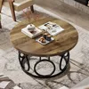 A round coffee table with a wood-grain top and a black metal base, placed on a rug in a living room setting. There are books, a vase with dried flowers, and cups with cookies on the table. Podgląd zdjęcia produktu