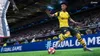 A soccer player in a yellow and black uniform kicks a soccer ball on a green field in a stadium. Produktbild-Vorschau